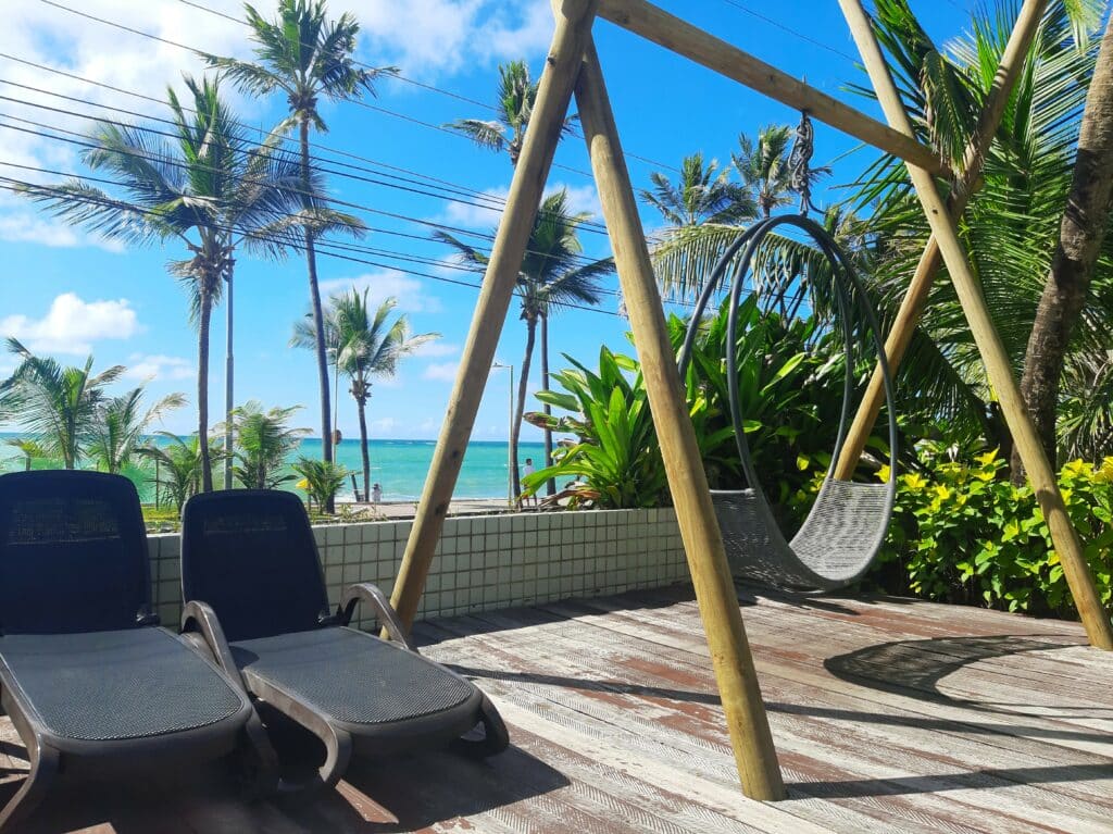 Poltronas e balanço num deck com folhas verdes ao lado (direito) e vista do mar ao fundo com coqueiros balançando com o vento