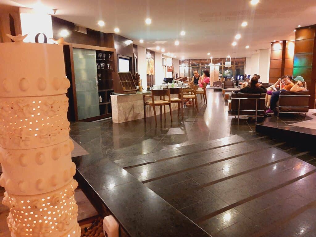 Bar localizado no lobby do Hotel Ritz Lagoa da Anta, com um balcão com cadeiras em volta e algumas pessoas sentadas, e algumas poltronas com hóspedes sentados conversando