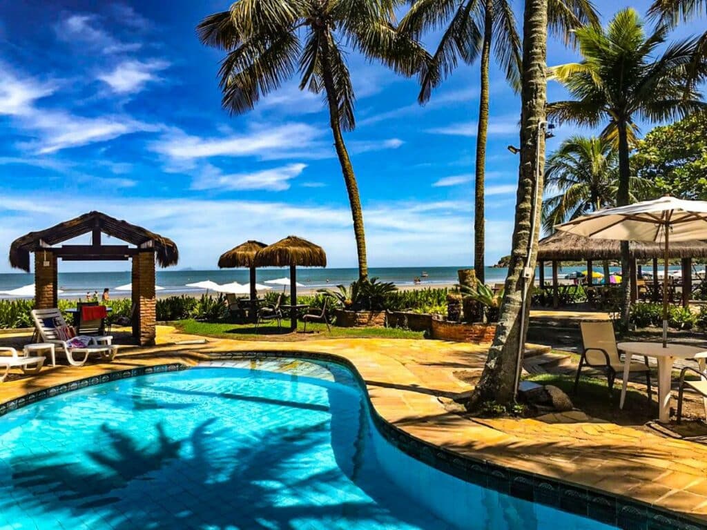 Piscina do Barequeçaba Praia Hotel cercada por mesinhas com cadeiras e coqueiros. Ao fundo é possível ver a praia.
