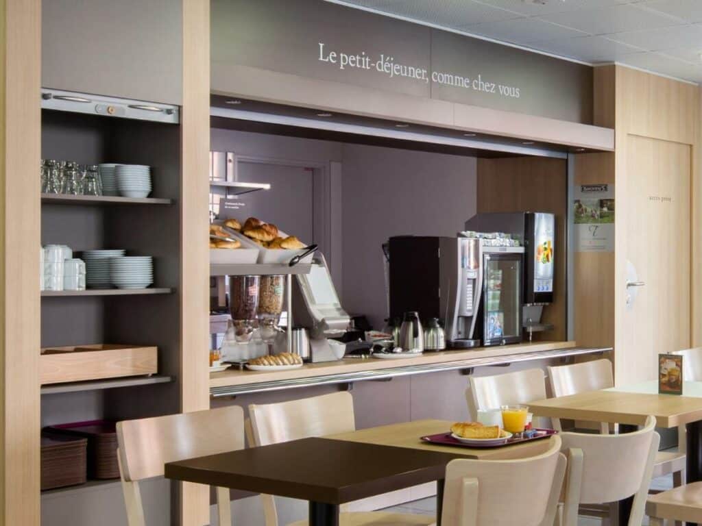 Área de refeições do B&B HOTEL Orly Chevilly Marché International com mesas, cadeiras e bancadas com utensílios de cozinha, itens de café da manhã e máquinas de café e de suco.