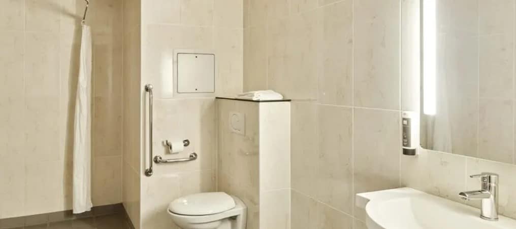 Banheiro adaptado para PCDs no B&B HOTEL Orly Chevilly Marché International. Ao lado esquerdo fica a área de banho com cortina. No centro está a privada com barras de apoio, e no canto direito fica a pia com espelho iluminado.