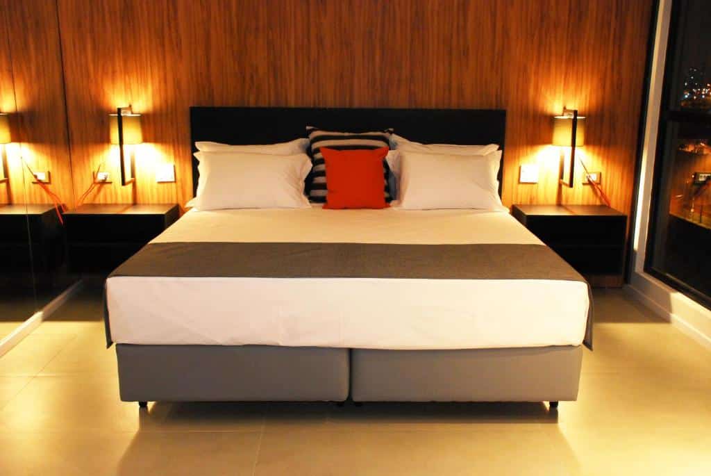 Foto do quarto no Best Guest Hotel Expo Anhembi. Vemos uma cama box de casal com almofadas. Em seus dois lados há uma mesa de cabeceira com abajur. É uma indicação de hotéis perto da Dutra.