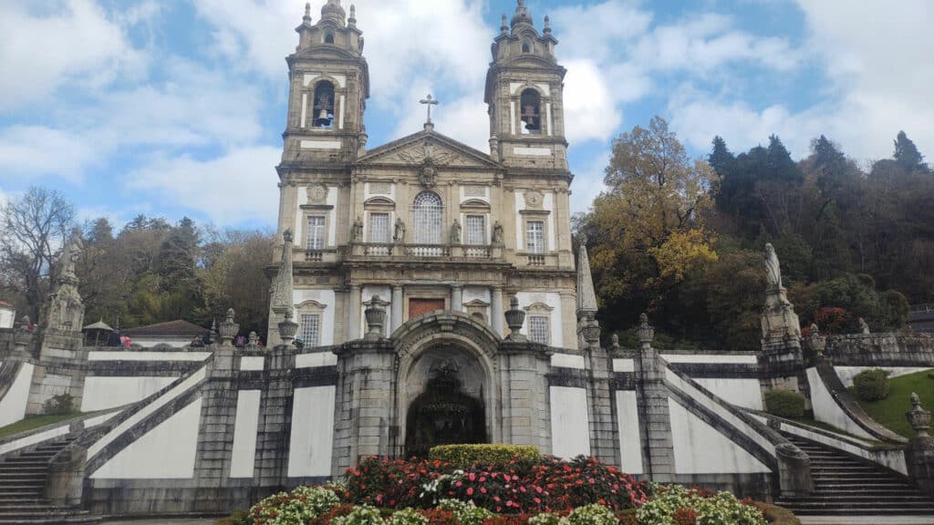 Santuário do Bom Jesus do Monte em Braga, uma igreja barroca com torres sineiras gêmeas e uma grande escadaria, cercada por vegetação e flores sob um céu parcialmente nublado.