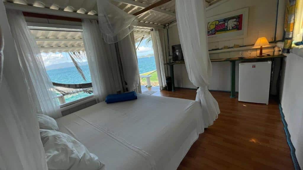 Quarto da Cabana Do Mar. No lado esquerdo está a cama de casal, na frente da cama há uma cômoda com TV, abajur e um frigobar e ao fundo a janela e a porta possuem vista para o mar.