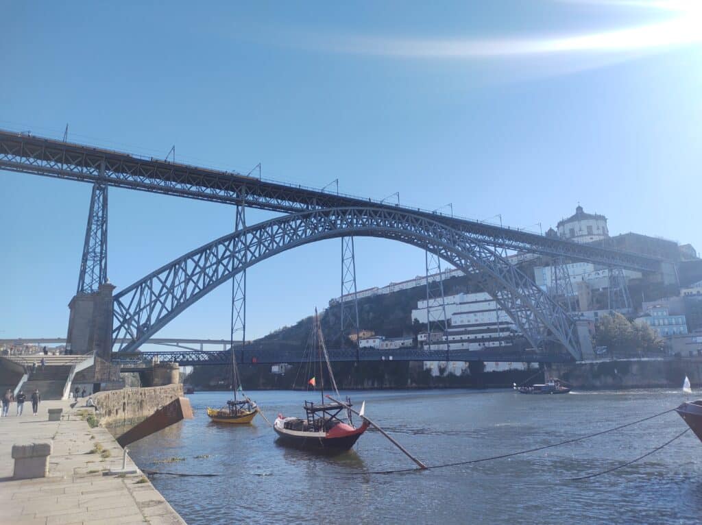 Ponte D. Luís I durante o dia com barcos do lado direito da imagem dentro do rio e ao fundo a ponte.