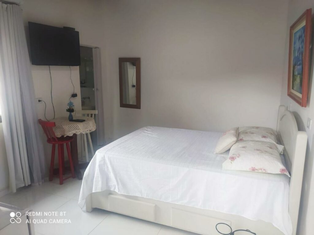Quarto do Cantinho de Berê, um dos airbnb em Barequeçaba. A cama de casal está encostada na parede direita e encara uma mesinha com crochê e duas cadeiras. Na parede acima fica uma TV.
