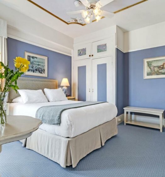Quarto do Cornell Hotel de France com uma mesa do lado esquerdo da imagem a frente com um vaso de flor ao fundo uma cama de casal e do lado esquerdo da cama uma cômoda com luminária. Representa hotéis em San Francisco.