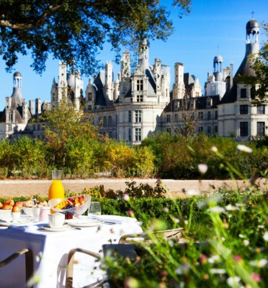Frente do Relais de Chambord – Small Luxury Hotels of the World durante o dia com mesa de café a frente no meio do jardim ao fundo o castelo. Representa hotéis no Vale do Loire.