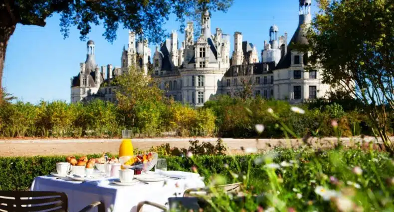 Frente do Relais de Chambord – Small Luxury Hotels of the World durante o dia com mesa de café a frente no meio do jardim ao fundo o castelo. Representa hotéis no Vale do Loire.