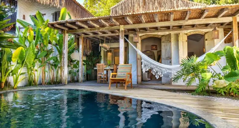Frente da Vila Rudá Trancoso durante o dia com piscina a frente ao fundo uma rede presa na parede da casa. Representa airbnb em Trancoso.