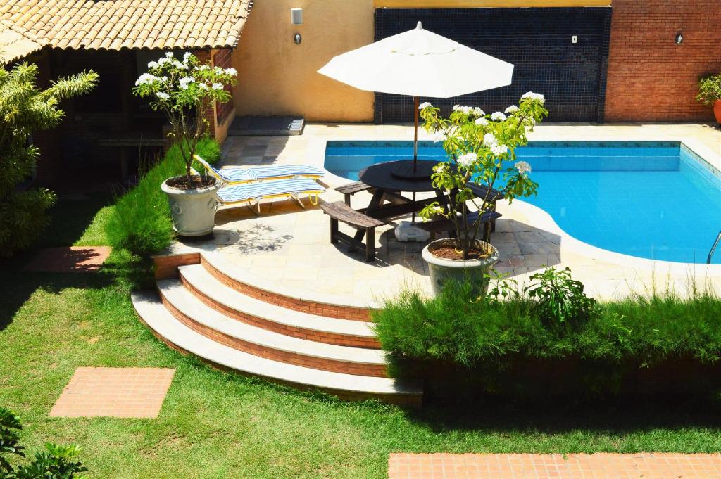 Área externa da Casa Atalaia. Um jardim, no meio uma área com cadeiras de sol, mesa de madeira com guarda-sol e uma piscina.