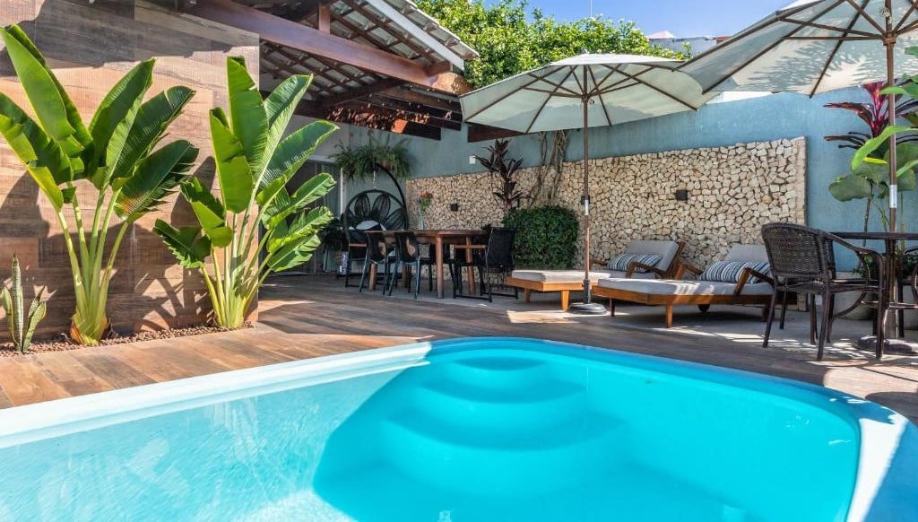 Área externa da Casa Caju para ilustrar post sobre airbnb em Aracaju. Na frente uma piscina, no fundo cadeiras de tomar sol, mesa e cadeiras com guarda-sol.