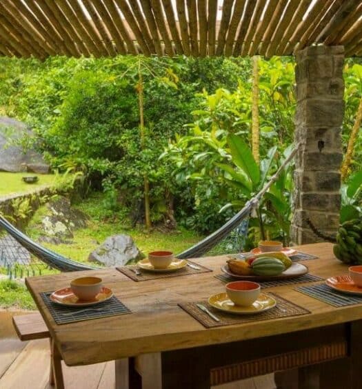 Área externa da Casa das Pedras para ilustrar post sobre airbnb em Ubatumirim. Uma mesa de café da manhã posta, no fundo uma rede e a vista do jardim.