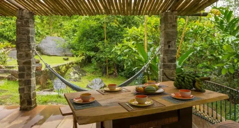 Área externa da Casa das Pedras para ilustrar post sobre airbnb em Ubatumirim. Uma mesa de café da manhã posta, no fundo uma rede e a vista do jardim.
