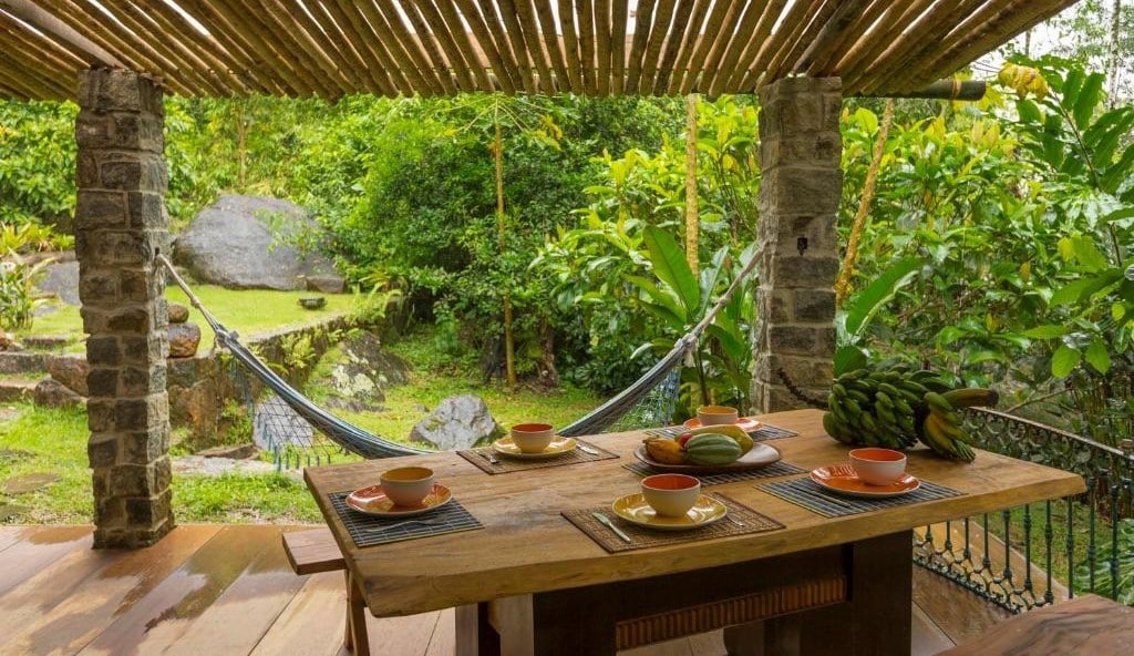 Área externa da Casa das Pedras para ilustrar post sobre airbnb em Ubatumirim. Uma mesa de café da manhã posta, no fundo uma rede e a vista do jardim.