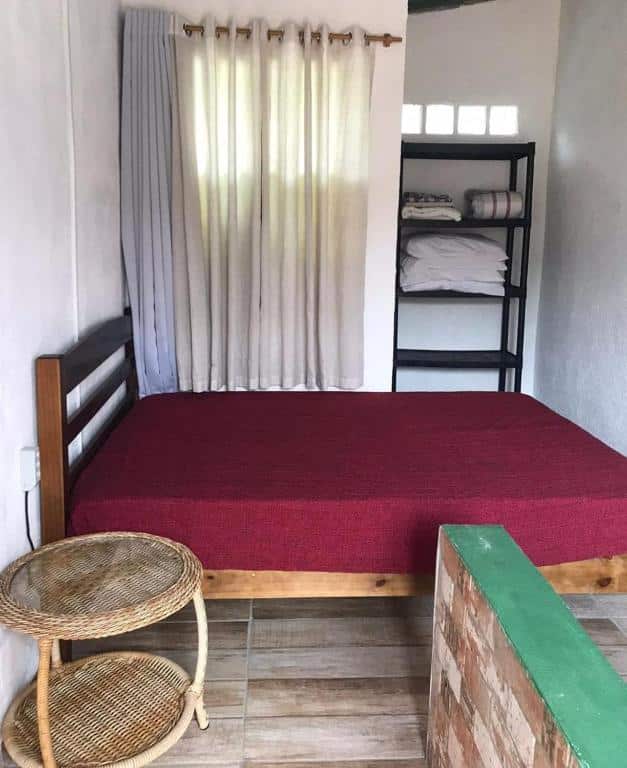 Quarto da Casa dos Passarinhos. Na frente um mesinha e atrás a cama de casal, com uma estante e a cortina fechada.