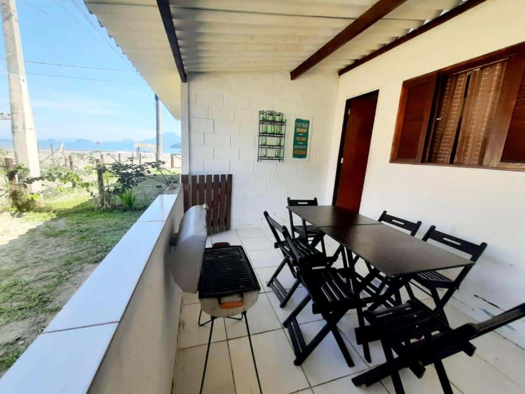 Área externa da Casa 45 Estaleiro. Uma mesa de madeira com cadeiras na varanda do lado direito, no meio uma churrasqueira e um muro médio. Do lado esquerdo a areia da praia.