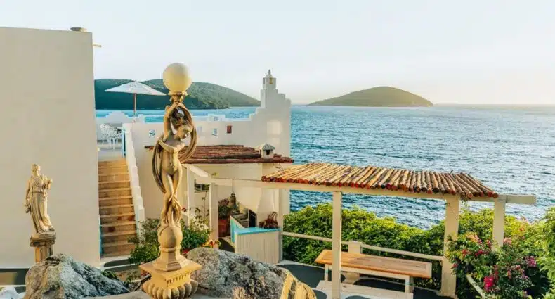 Vista à beira-mar da Casa Grega, um dos Airbnb em Arraial do Cabo, com uma pérgula, estátuas e colinas verdes do outro lado da água sob um céu claro.