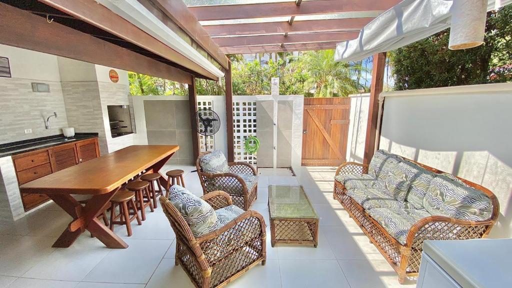 Área de churrasco do airbnb "Casa- Maresias 3 minutos da praia". Do lado esquerdo há uma churrasqueira e uma pia lado a lado, e de frente para elas uma mesa de madeira de vários lugares. Do lado direito há um sofá e duas poltronas ao redor de uma mesinha no meio. Ap fundo há um veentilador, uma mangueira e a porta para a rua.