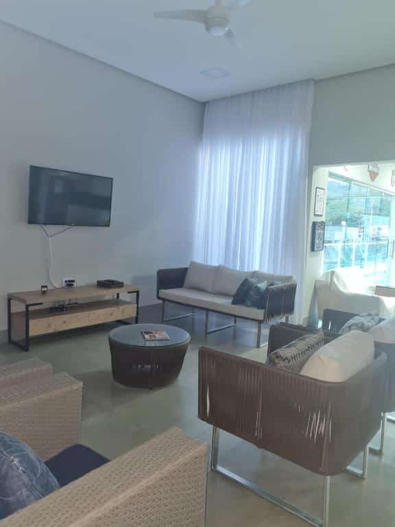 Sala de estar da Casa Tô D`Boa Capitólio que mostra quatro poltronas, um sofá, uma mesinha redonda no meio, uma mesa encostada na parede e em cima uma tv. Atrás do sofá tem uma cortina e uma parte de vidro. Imagem ilustrando post Airbnb em Capitólio.
