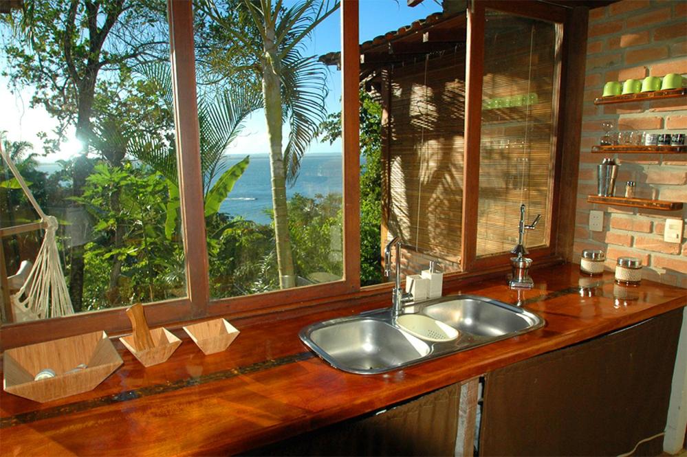 Cozinha da Casa Vista al Mar. Do lado direito prateleiras com copos e xícaras. No meio uma pia com uma janela de vidro e vista para o mar.