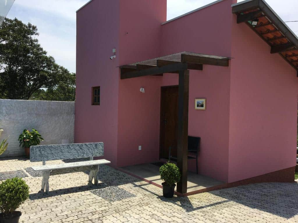 Parte externa do Chalé Mantiqueira que mostra uma casa cor de rosa com uma porta de madeira, uma janela, uma cadeira, uma cobertura de madeira com vidro e um banco na frente do chalé. Imagem ilustrando post Airbnb em Cunha.