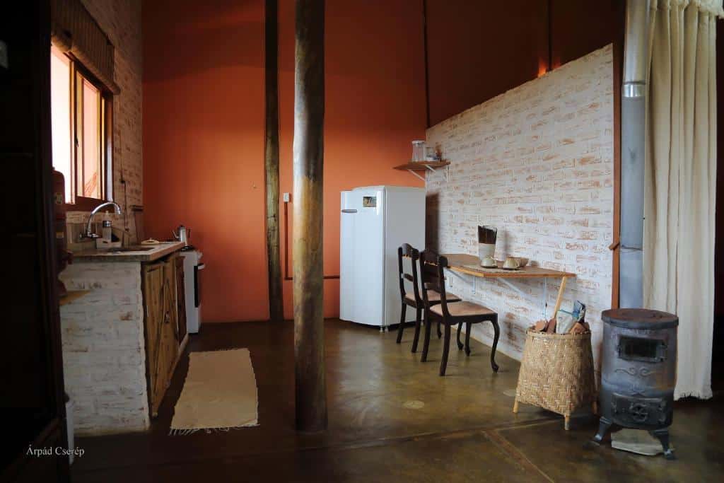 Parte externa do Chalés Samadhi que mostra a cozinha com com duas pilastras de madeira no meio e doa lado esquerdo a pia com um armário, um fogão e uma janela. Já do lado direito tem uma geladeira, uma mesa na parede com duas cadeiras, um cesto e outro objeto de ferro. Imagem ilustrando post Airbnb em Cunha.