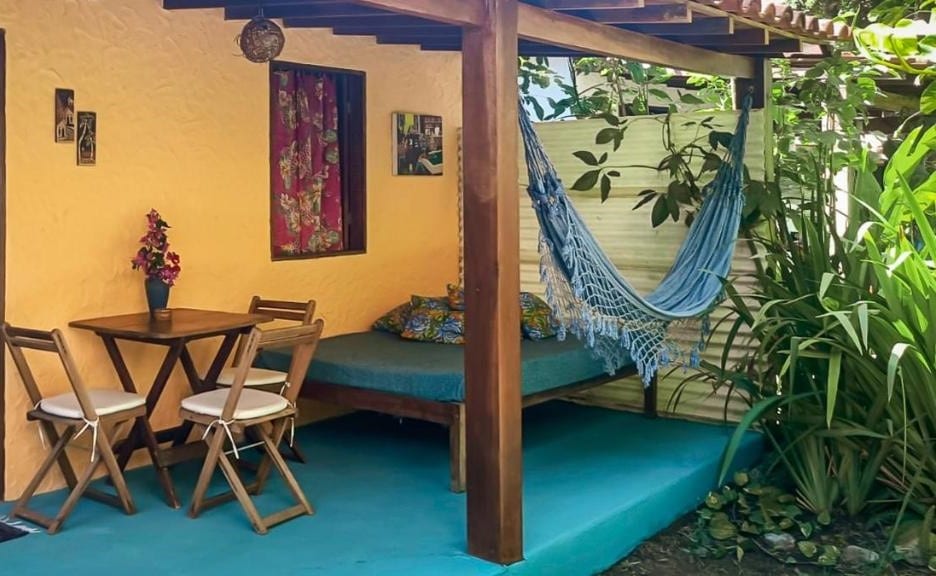 Área externa do Chalés Vila D'Ajuda. uma mesa pequena de três lugares está ao lado de uma cama com almofadas e uma rede. Todos eles estão debaixo de um pequeno telhado de madeira.
