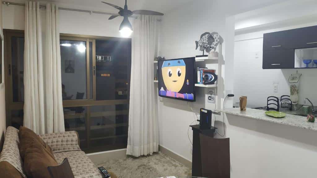 Sala de estar do Concept Tower Praia Apart. Do lado direito um balcão, atrás a cozinha. Uma televisão e umas prateleiras. No meio um ventilador de teto, do lado esquerdo um sofá. No fundo uma parede de vidro.