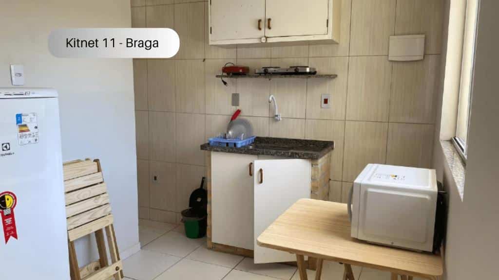 Cozinha do Cabo Frio - Braga - Kitnets - Aluguel Econômico. No lado esquerdo está a geladeira e um corredor, no lado direito há uma mesa com um microondas em cima dela e no fundo está a pia com uma prateleira e um armário logo acima.