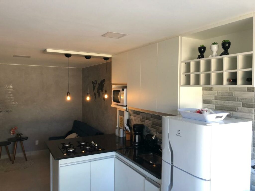 Cozinha do Paraíso na Ilha da Gigóia com geladeira do lado direito da imagem, do lado da geladeira u a pia e o fogão e ao fundo  a sala de estar com sofá.