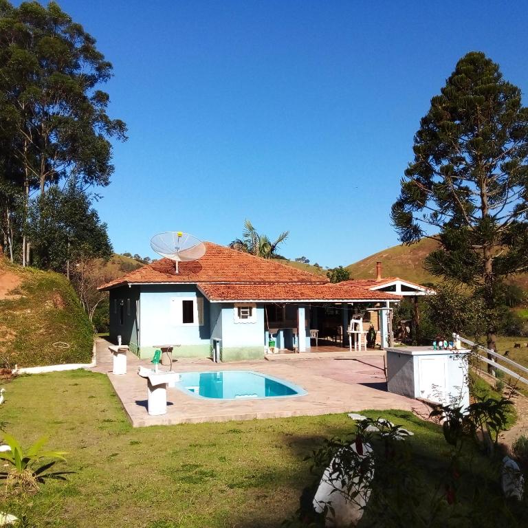 Parte externa do Estalagem Sitio Colibri que mostra a casa em tons de azul com algumas janelas, uma antena e um telhados com telhas cor terra. Na frente da casa tem uma piscina e em volta um amplo gramado verde e várias árvores em volta durante o dia. Imagem ilustrando post Airbnb em Cunha.