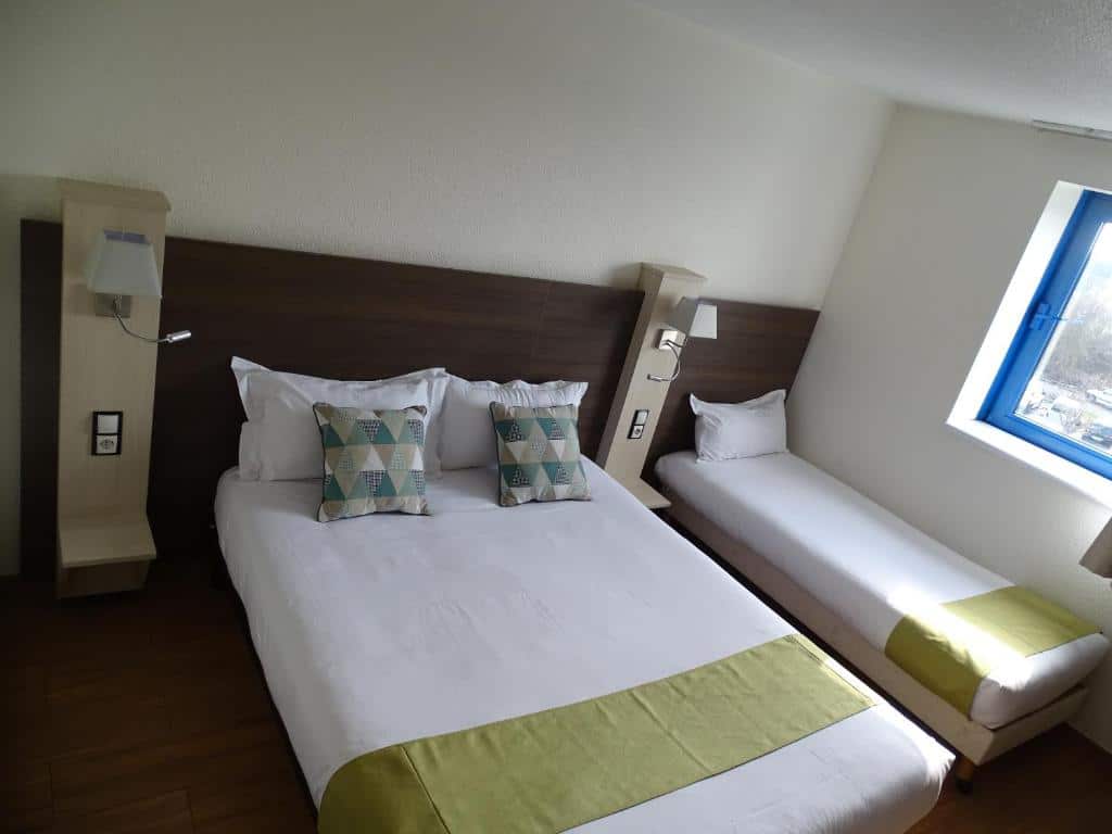 Visão de cima de um quarto do Euro Hôtel Airport Orly Rungis, um dos hotéis perto do Aeroporto de Orly. Uma cama de solteiro e uma de casal tem uma mesinha de cabeceira entre si e luminárias dos dois lados, assim como uma janela na parede direita.