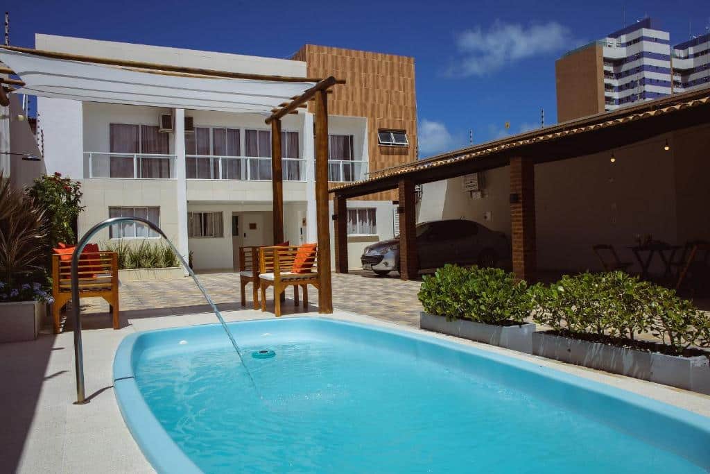 Área externa do Flat com 2 quartos. Uma piscina na frente com cascata, no fundo cadeiras, um carro estacionado e a casa.