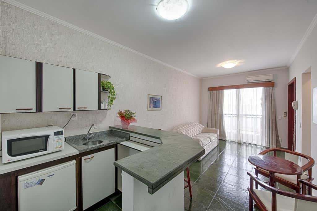 Flat Itararé Tower. Do lado esquerdo uma cozinha com armários, pia, micro-ondas, frigobar e um balcão. Do lado direito uma mesa redonda pequena com dois bancos. No fundo a sala de estar com um ar-condicionado.