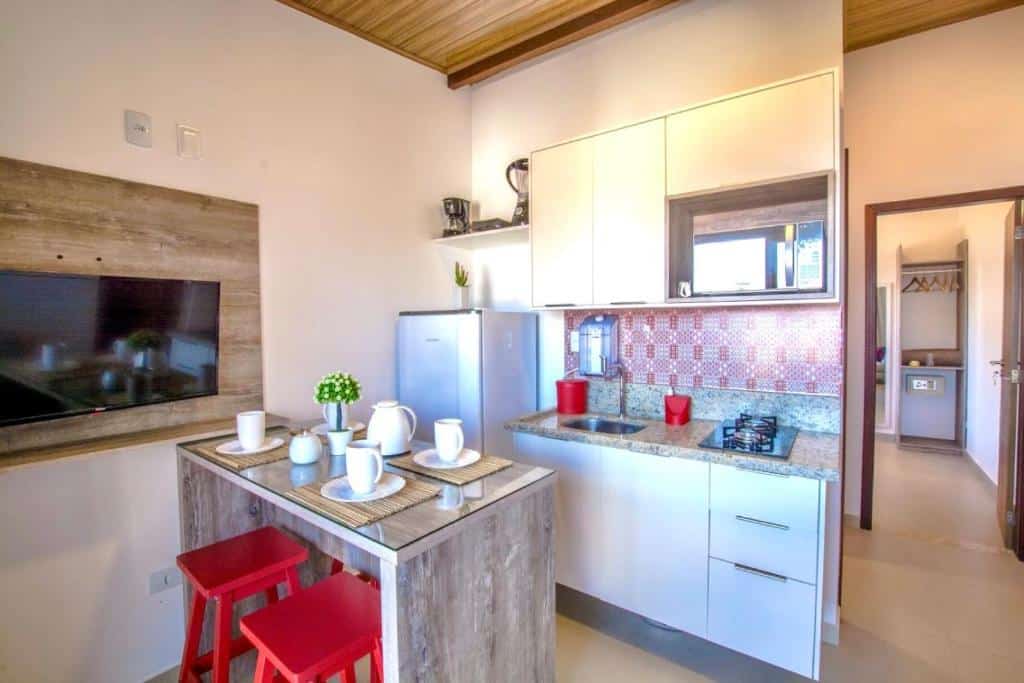 Cozinha do Flats Morro de São Paulo. Do lado esquerdo uma televisão, uma banqueta posta com bancos. Atrás uma mini cozinha. No fundo, no lado direito, o corredor.