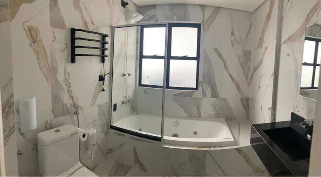 Banheiro do Flats São Vicente. Do lado direito a pia e de frente o vaso sanitário. No fundo um box com uma banheira de hidro e uma janela.