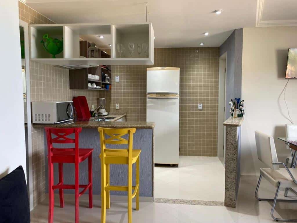Cozinha do airbnb Garatucaia. No lado esquerdo há um balcão com um microondas em cima, duas banquetas atrás do balcão, uma pia, um armário em cima da pia e uma geladeira ao lado. Já no lado direito é possível ver uma porta, outro balcão e uma parte de uma mesa com cadeiras em volta.