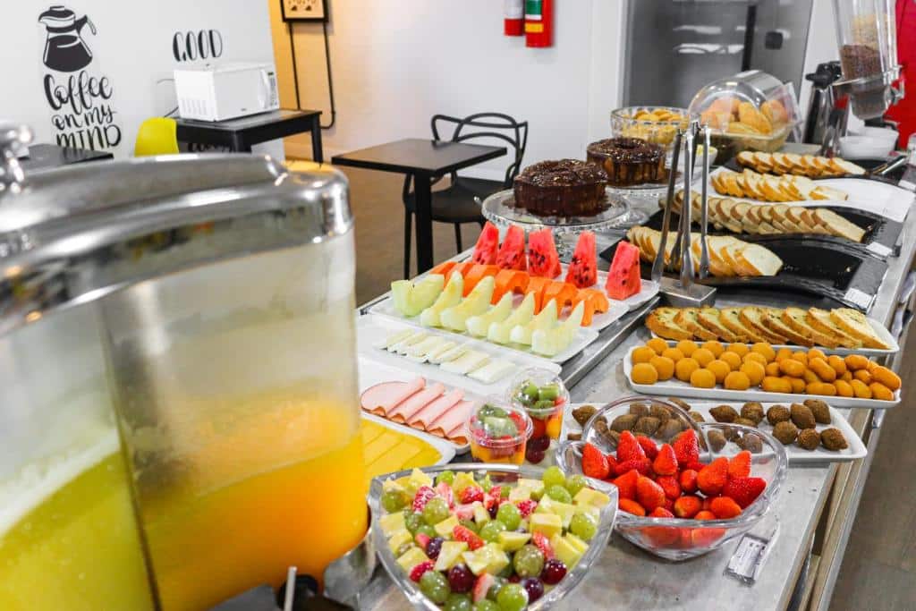 Buffet de café da manhã do GRAAL INN ITATIAIA. A bancada possui diversas frutas, frios, salgadinhos, pães e bolos.