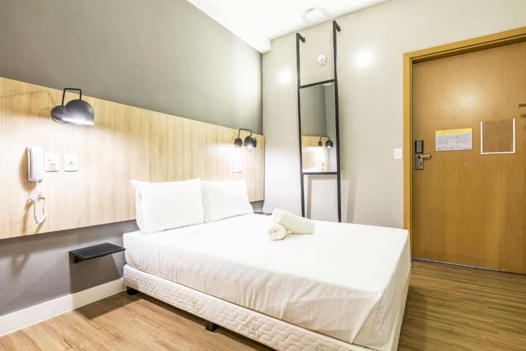 Quarto espaçoso do GRAAL INN ITATIAIA. Uma cama de casal está na esquerda, no centro, e atrás há um painel com abajures e tomadas. Do lado direito da cama há um espelho e a porta de entrada para o quarto. É uma indicação de hotéis perto da Dutra.