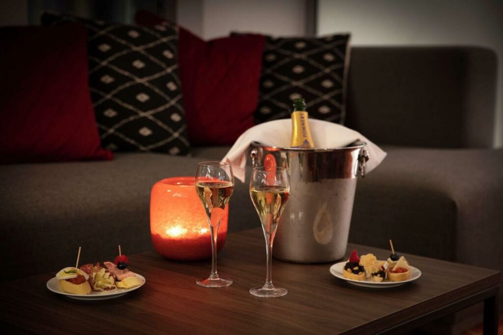 Mesa de madeira com um balde com champanhe, taças cheias do líquido, pratinhos de aperitivos e uma vela iluminando o ambiente. Há um sofá com almofadas logo atrás.