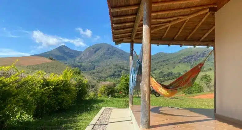 Parte da Hospedagem Colinas que mostra ao lado direito duas redes para descansar embaixo de uma varanda de pilastras de madeira e telhado de telhas. Do lado esquerdo tem algumas árvores e ao fundo a paisagem de montanhas durante o dia. Imagem ilustrando post Airbnb em Aiuruoca.