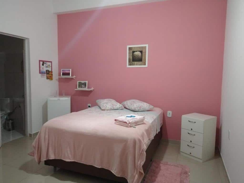 Quarto de 12m² da Hospedagem Elite térreo com uma cama de casal box ao centro do quarto. Do lado direito tem uma pequena cômoda e do lado esquerdo tem um frigobar, quadros na parede e uma porta aberta que dá acesso ao banheiro. Imagem ilustrando post Airbnb em Cunha. 