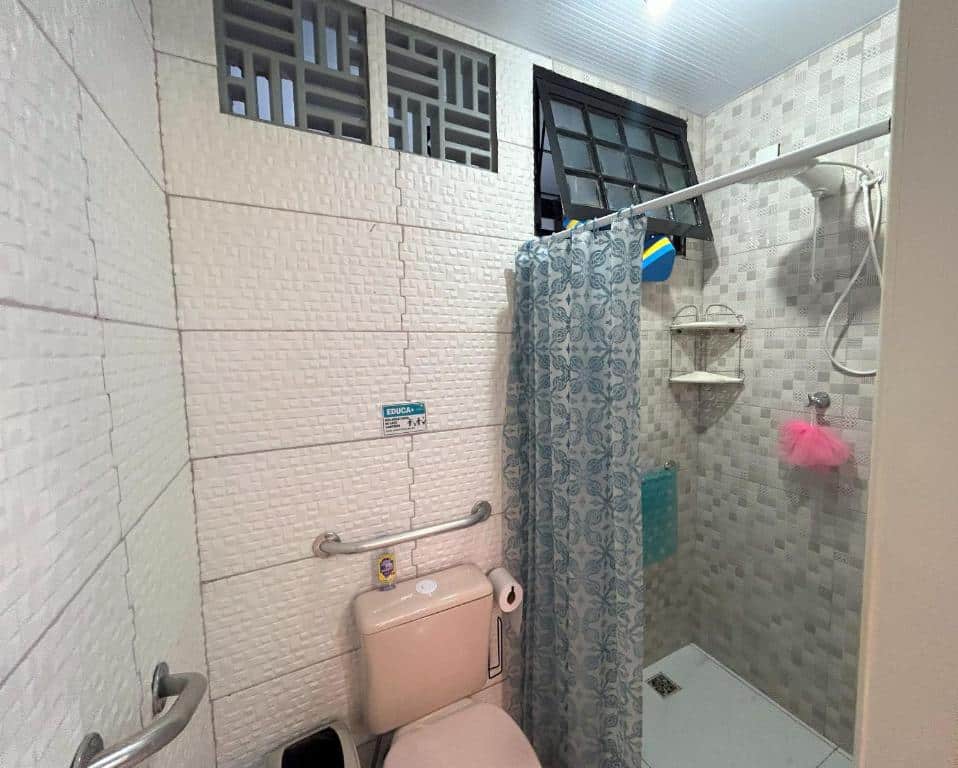 Imgem de um banheiro no Hostel Quintal de casa, com um vaso sanitário no centro com barras de apoio ao seu redor. Uma ducha está à direita da foto.