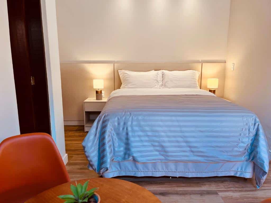 Quarto do Hotel do Parque - Cunha com 21m² e uma cama de casal com travesseiros e cobertores em cima e de cada lado tem uma mesinha com um abajur. Na frente da cama tem parte de uma mesa de madeira e uma cadeira aparecendo e uma porta do lado esquerdo do quarto. Imagem ilustrando post Airbnb em Cunha.