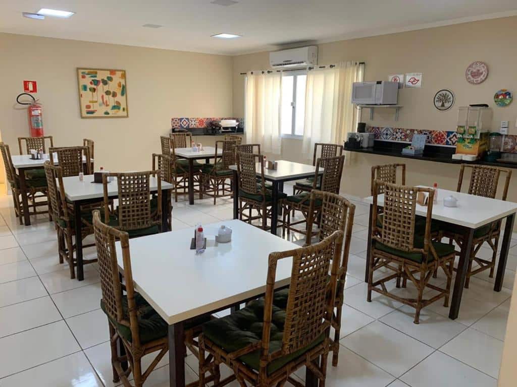 Refeitório do Hotel e Pousada Areia da Praia. Mesas com cadeiras no meio. Atrás um balcão com sucos, tostadeira, micro-ondas. Uma janela e em cima um ar-condicionado.