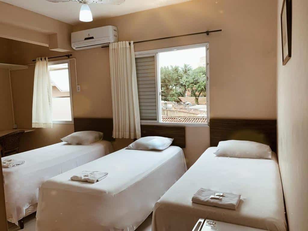 Quarto do Hotel e Pousada Areia da Praia. Três camas de solteiro com toalhas, travesseiros e itens de banho. No canto direito um frigobar, no fundo duas janelas e em cima um a-condicionado. Foto para ilustrar post sobre pousadas em São Vicente.
