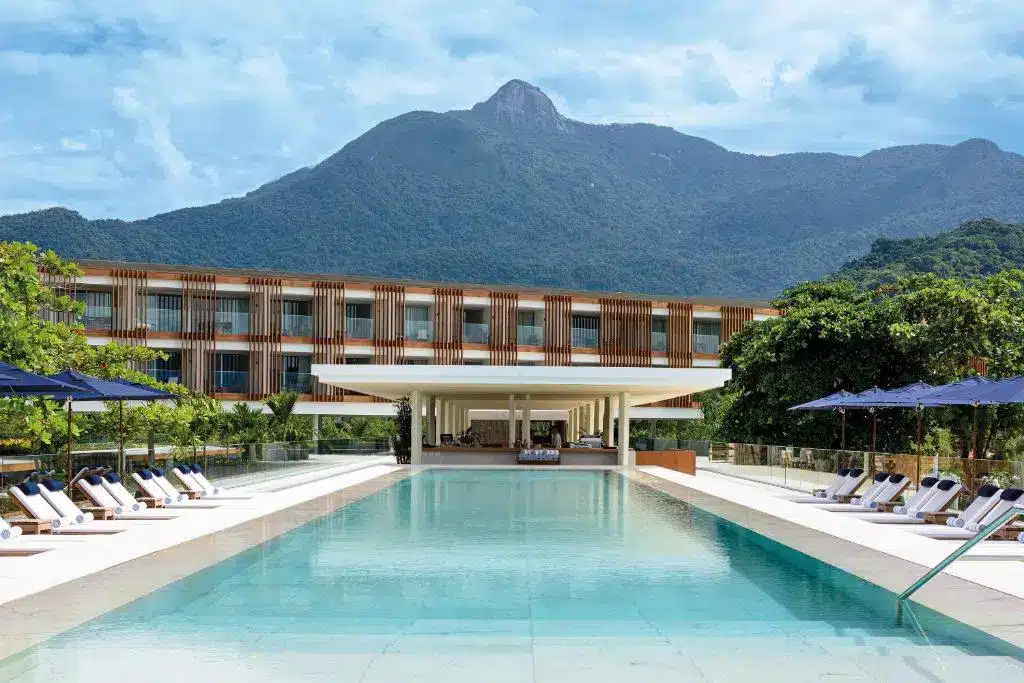 Piscina do Hotel Fasano Angra dos Reis. A piscina está centralizada, nos lados estão espreguiçadeiras e ao fundo está a estrutura do hotel com uma montanha atrás.