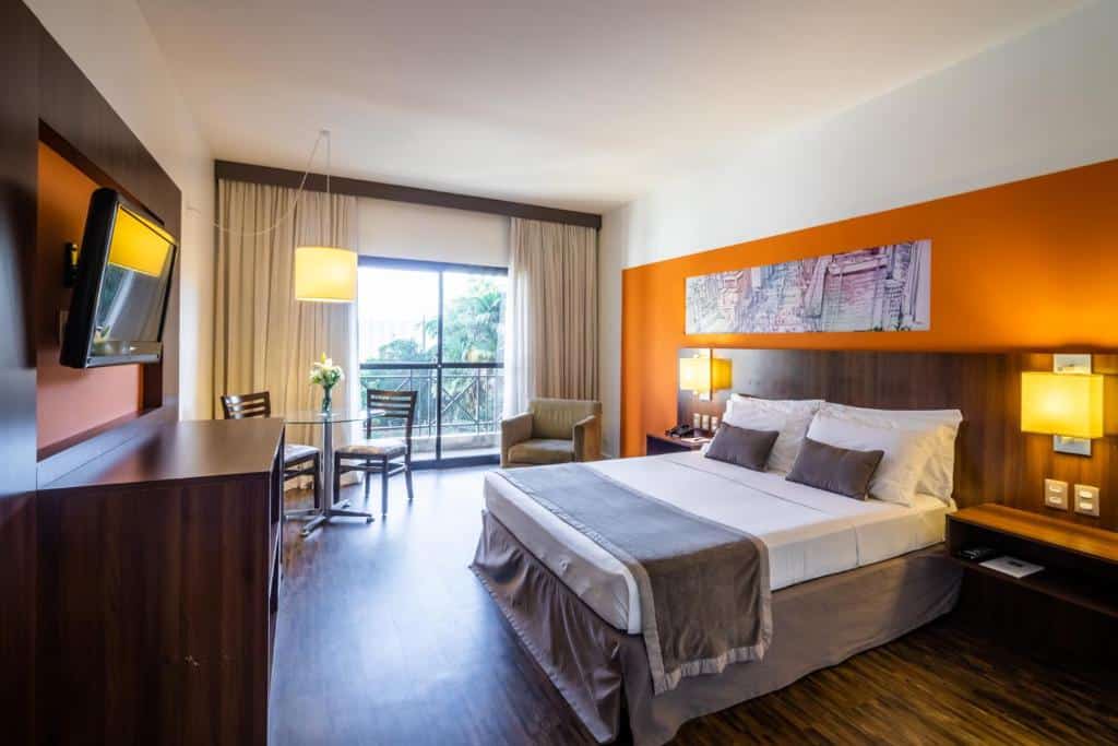 Quarto do Hotel Panamby Guarulhos. Há uma cama king-size à direita, atrás dela há uma cabeceira com abajures. Na esquerda da cama, no fundo da foto, há uma poltrona e uma mesa com duas cadeiras, além de uma porta com varanda. Na frente da cama há um painel de TV. É uma indicação de hotéis perto da Dutra.