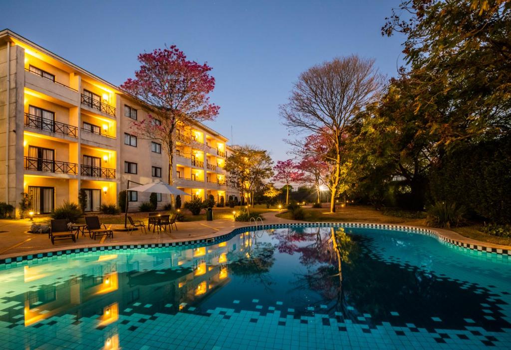 Área da piscina do Hotel Panamby Guarulhos. A piscina é enorme e reflete a luz vinda das acomodações do hotel à esquerda. Há várias árvores em volta.